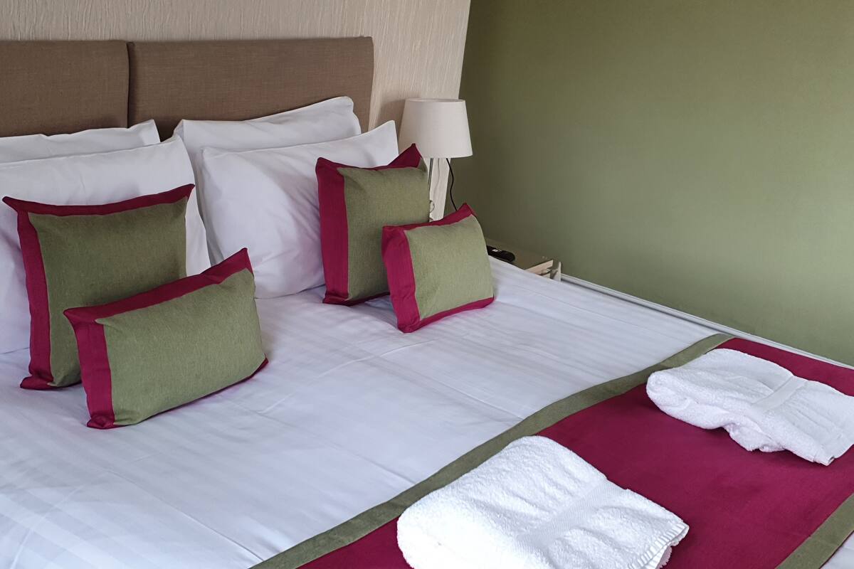 A neatly made bed with white linens, green and red accent pillows, a white bathrobe, and a red towel on a green bed runner.