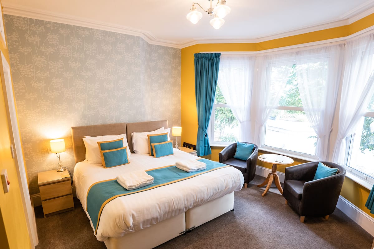 A hotel room with a large bed, two bedside tables, a small round table, two armchairs, and a window with curtains.