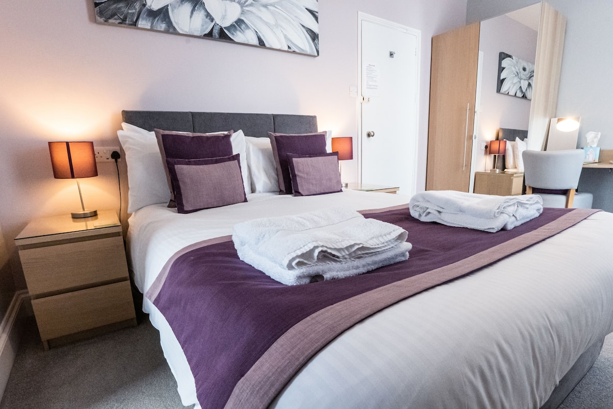 A neatly made bed with a purple throw blanket and folded towels, a nightstand with a lamp, and a door with a sign.