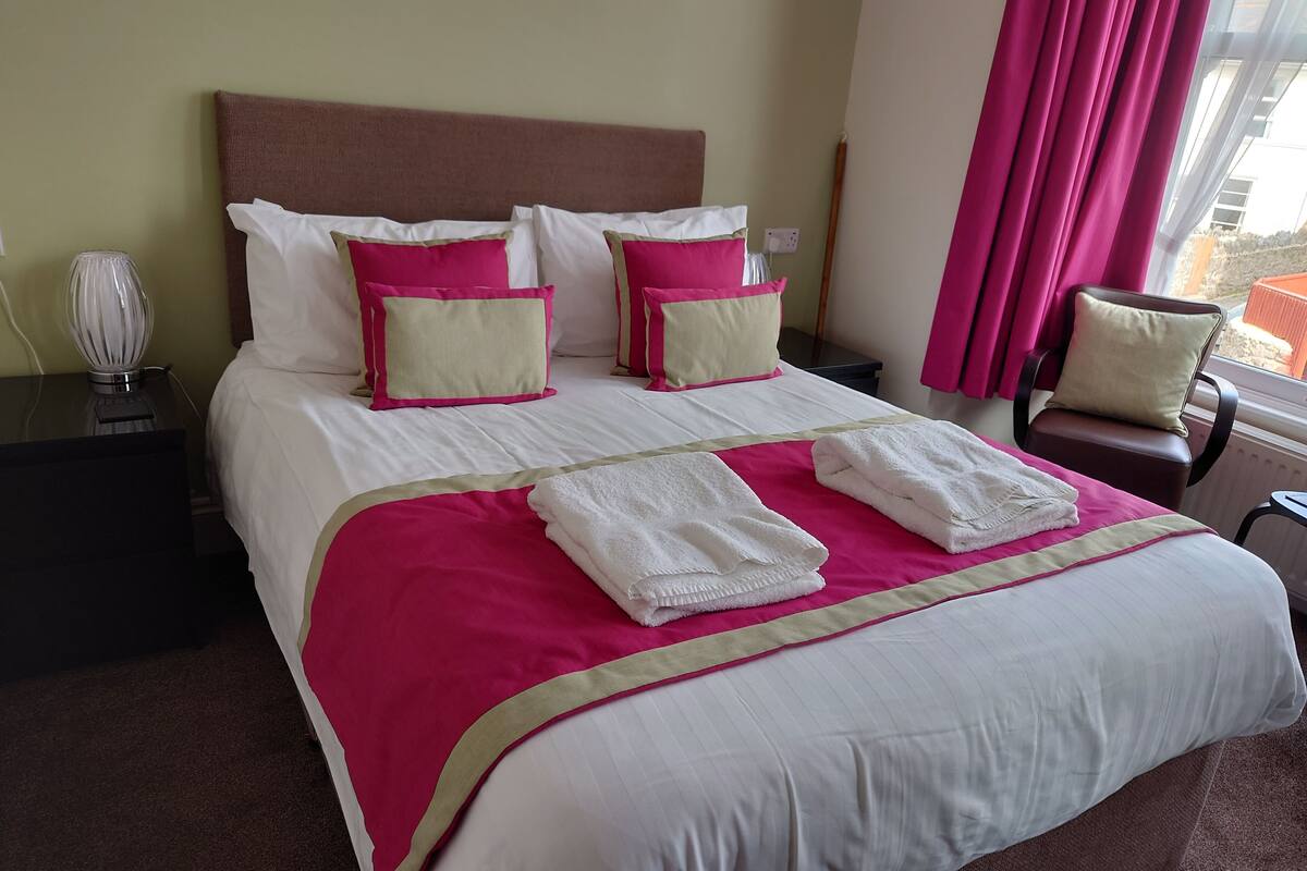 A neatly made bed with white and pink bedding, a bedside lamp, and a chair by the window.