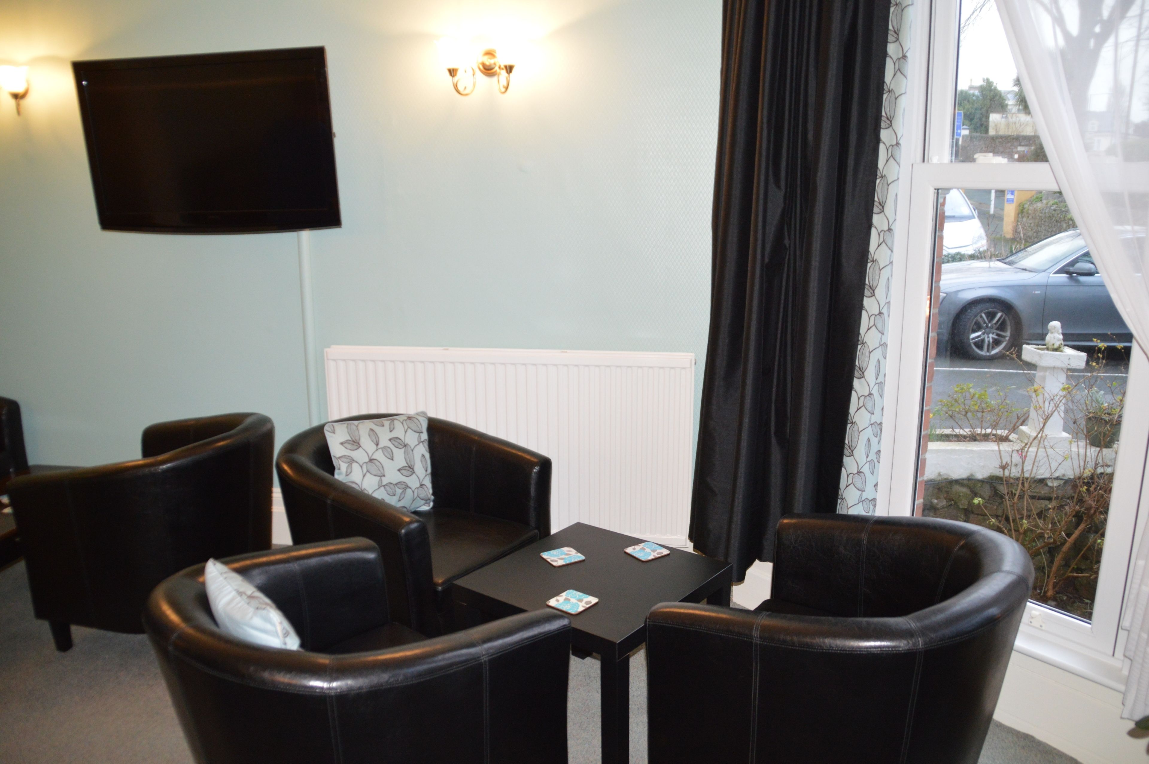 Une pièce comprenant quatre fauteuils en cuir noir, une petite table avec deux sets de table, un téléviseur à écran plat fixé au mur et une fenêtre donnant sur une voiture garée et sur la verdure à l’extérieur.