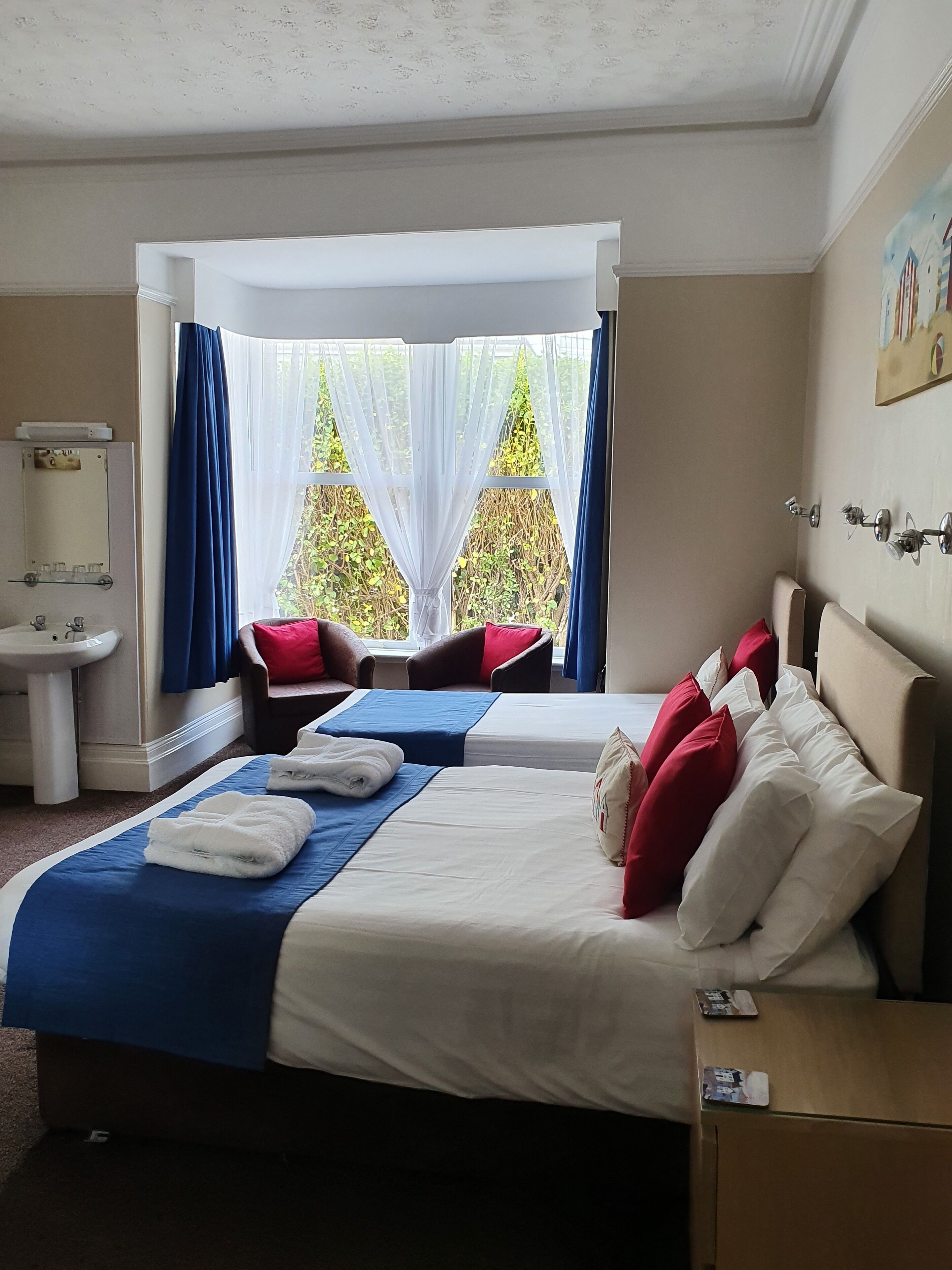 Habitación de hotel con dos camas, una zona de estar con una silla y una mesa, un lavamanos y una ventana con cortinas traslúcidas.