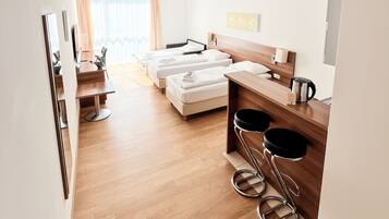 A hotel room with a wooden floor, a bed, a desk, two bar stools, and a coffee machine.