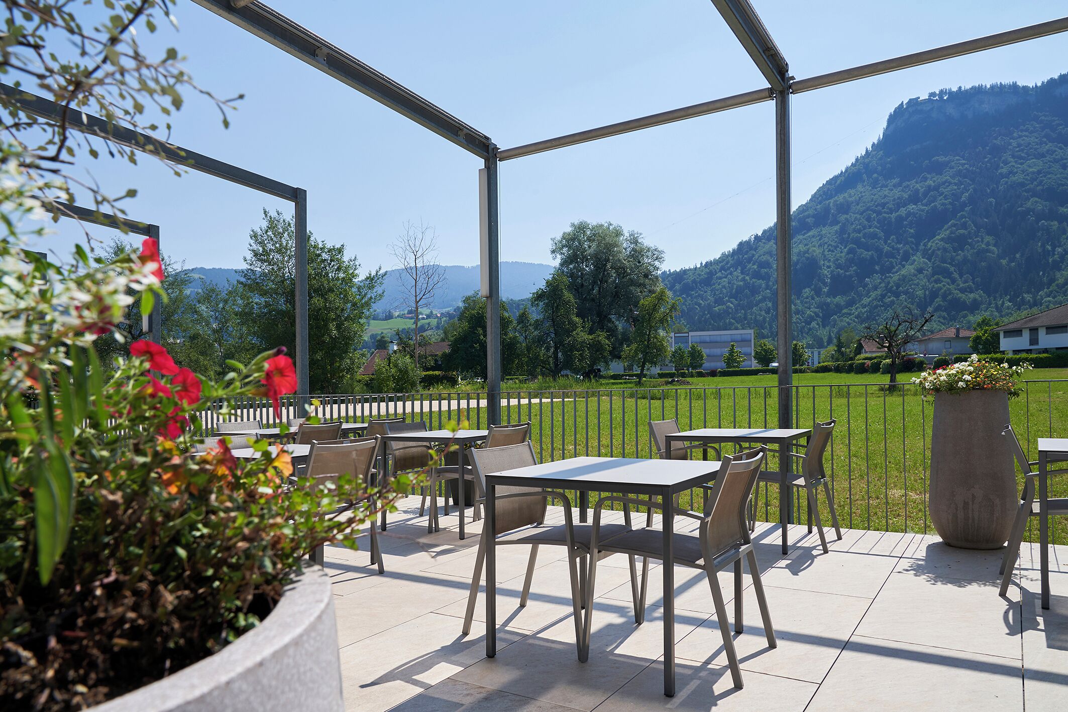 Een terras buiten met tafels en stoelen, een grote plantenbak met bloemen, en uitzicht op een groen landschap en heuvels.