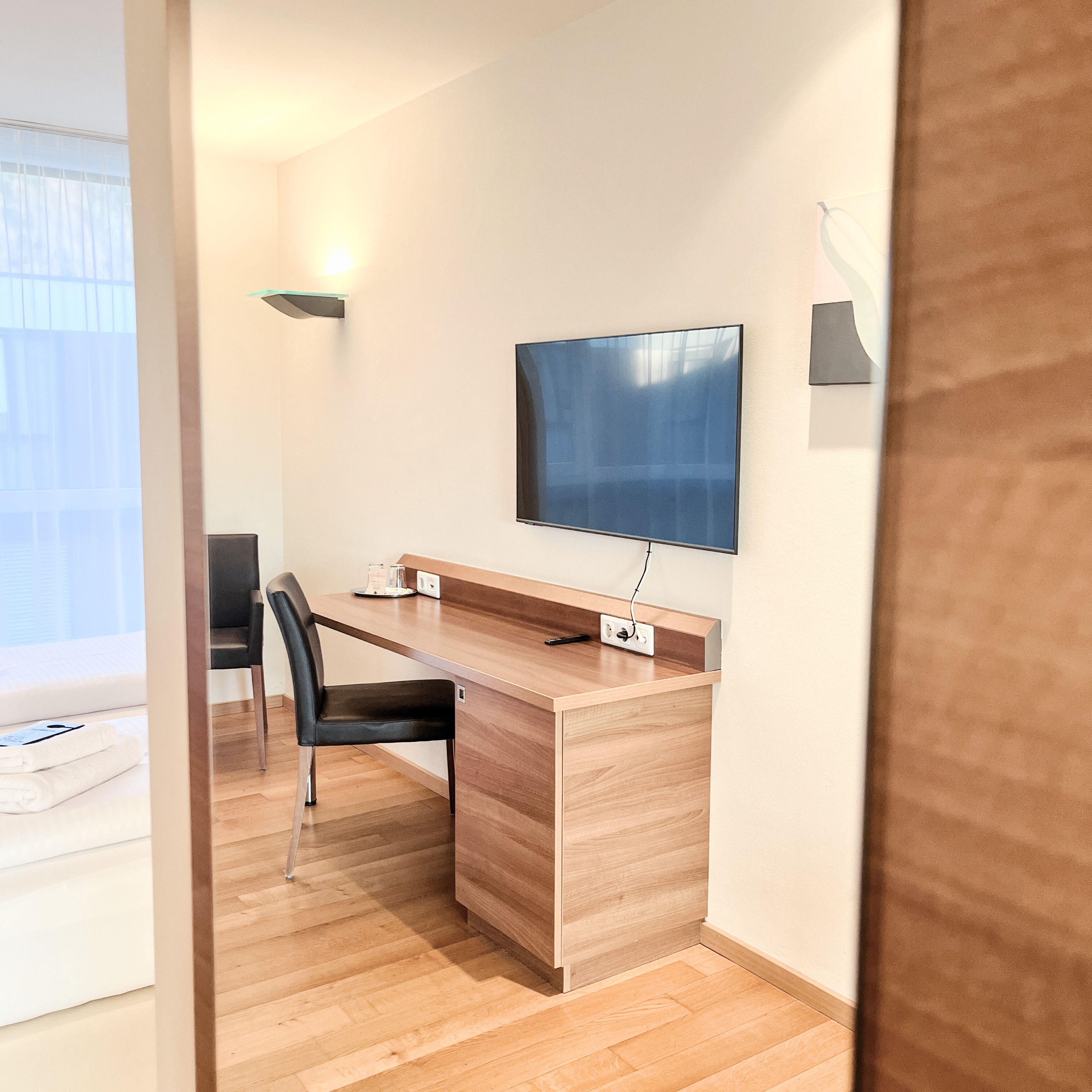 Een moderne kamer met een houten bureau, een zwarte stoel, een flatscreen televisie aan de muur en een klein bijzettafeltje.