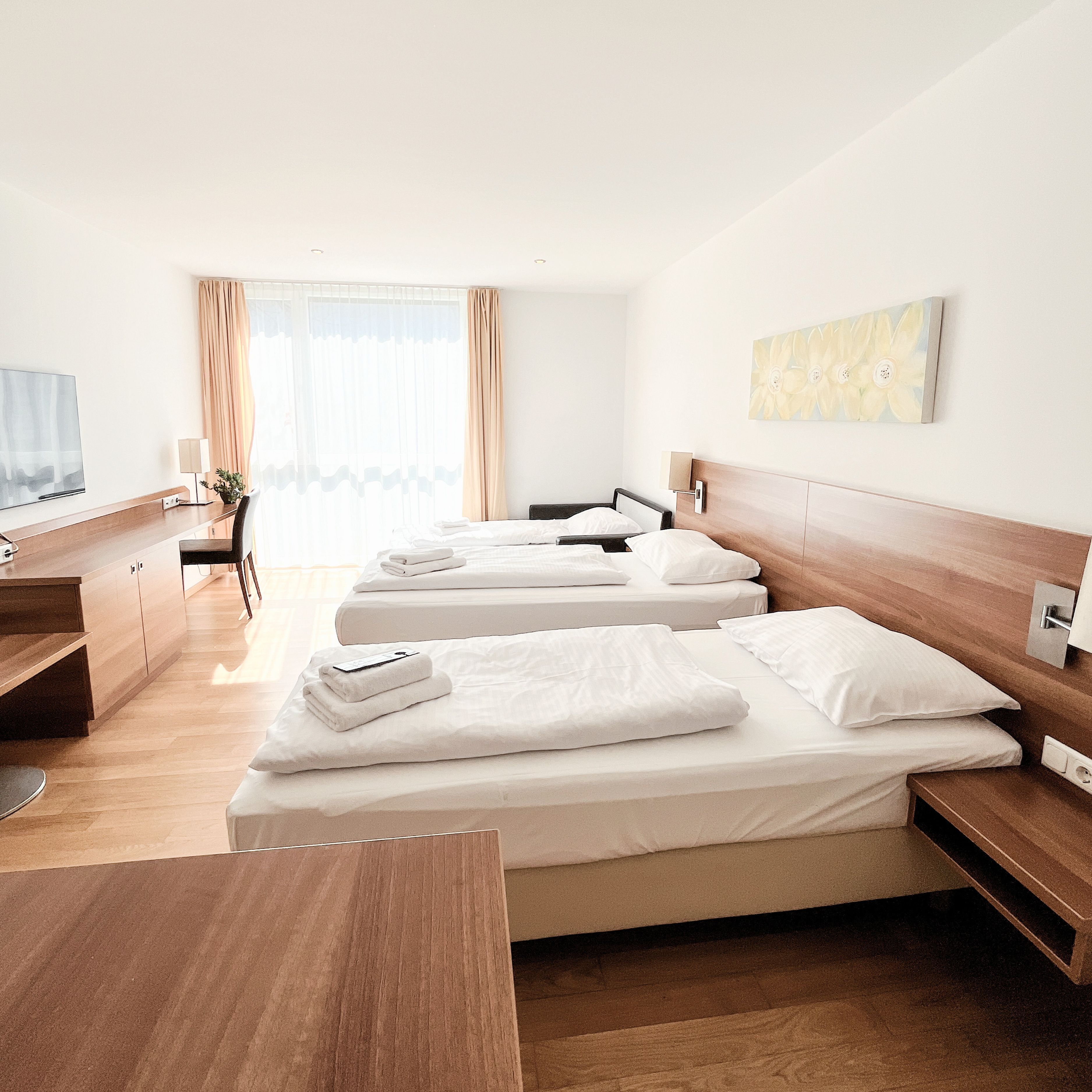 Habitación de hotel con tres camas, un escritorio, televisor y ventana con cortinas.