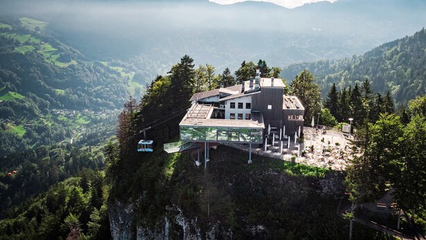 Un chalet de montagne doté d’un pont de verre, entouré de collines boisées et accessible par un téléphérique.