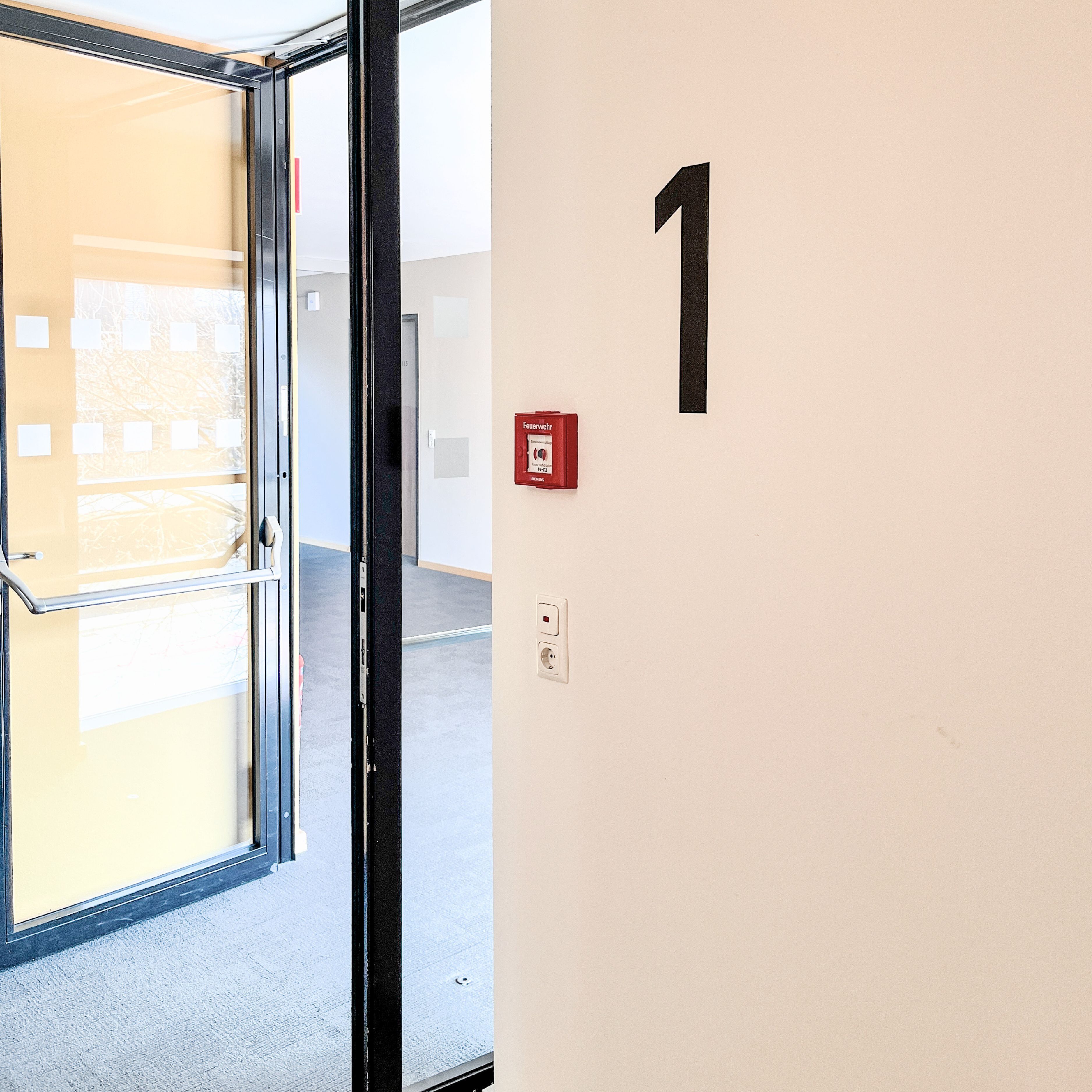 A door with a glass panel, a fire alarm, and a wall with the number 1.