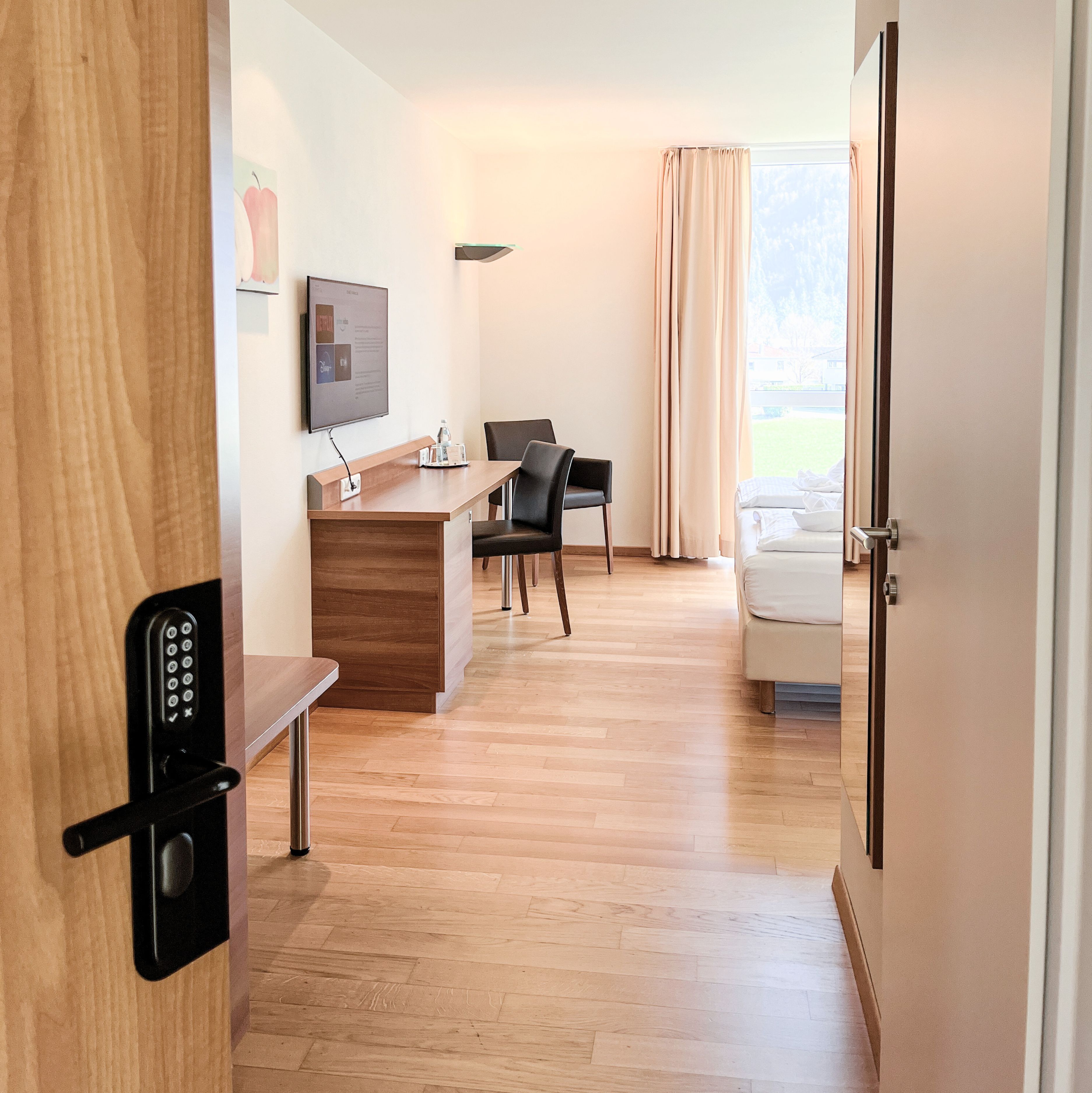 Ein Hotelzimmer mit Schreibtisch aus Holz, einem an der Wand befestigten Fernseher, einem schwarzen Stuhl, einem weißen Sofa und einer Tür mit Zahlenschloss.