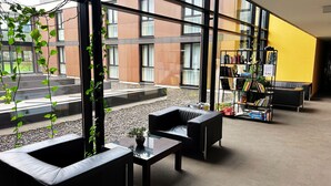 Un espace intérieur moderne avec des canapés en cuir noir, une table basse en verre, une bibliothèque et une vue sur un immeuble à travers de grandes fenêtres.