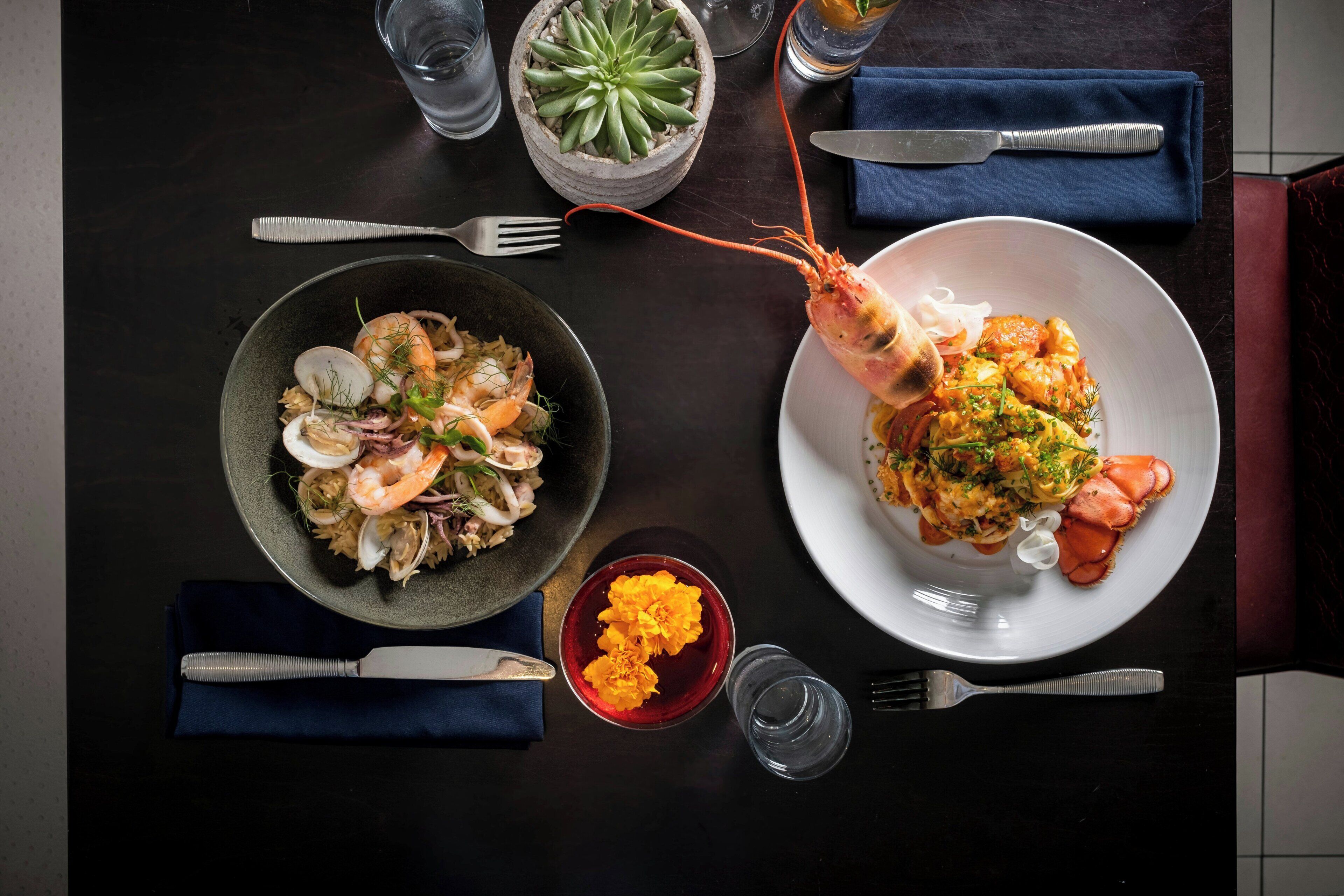 A dining table with two plates of seafood pasta, a glass of water, a small bowl of orange garnish, and a potted succulent plant.