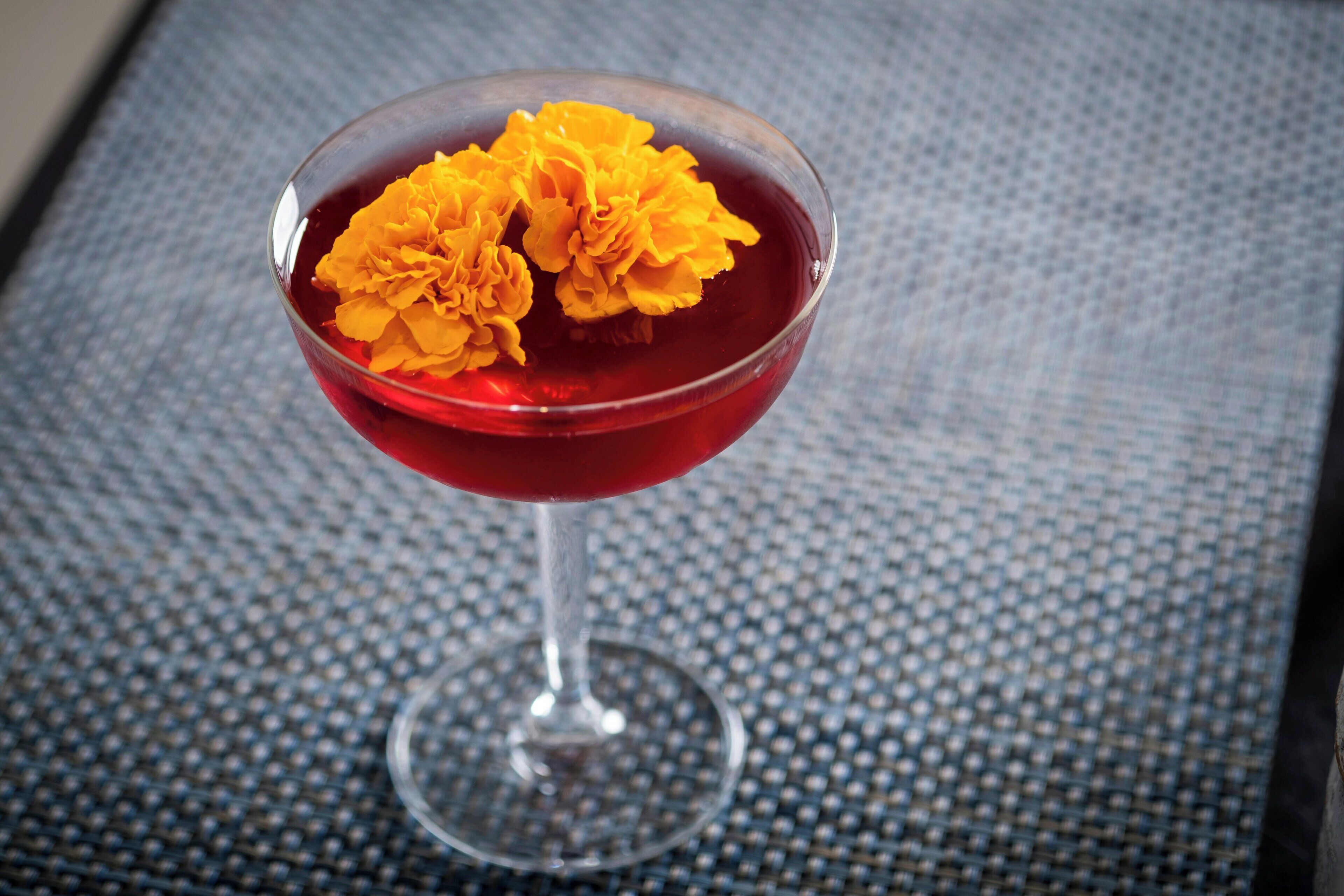 A cocktail glass with a red liquid and two orange flowers on a textured surface.