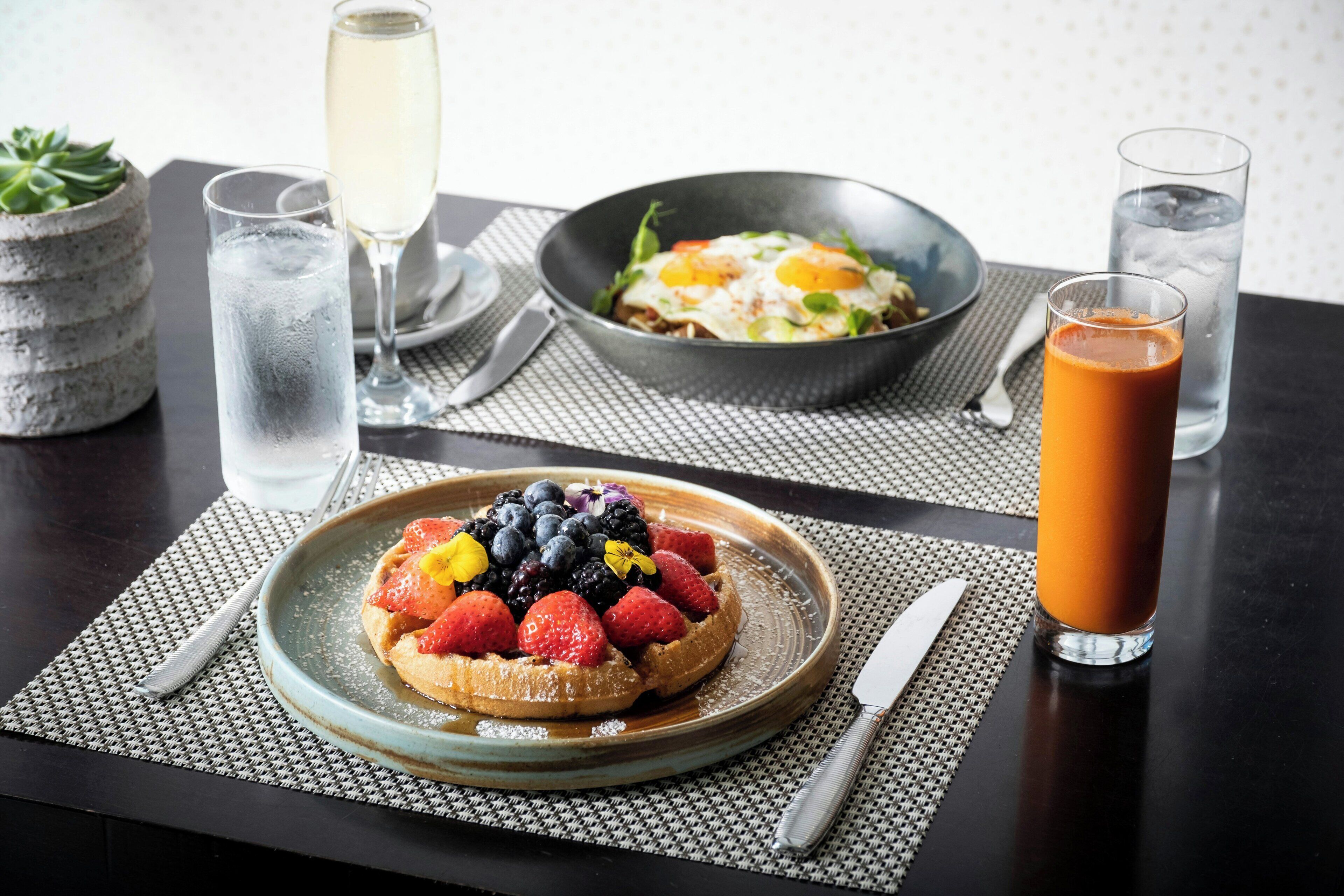 Une table mise pour le petit-déjeuner avec une assiette de tartines garnies de fruits, un bol d’œufs et un verre de jus d’orange.