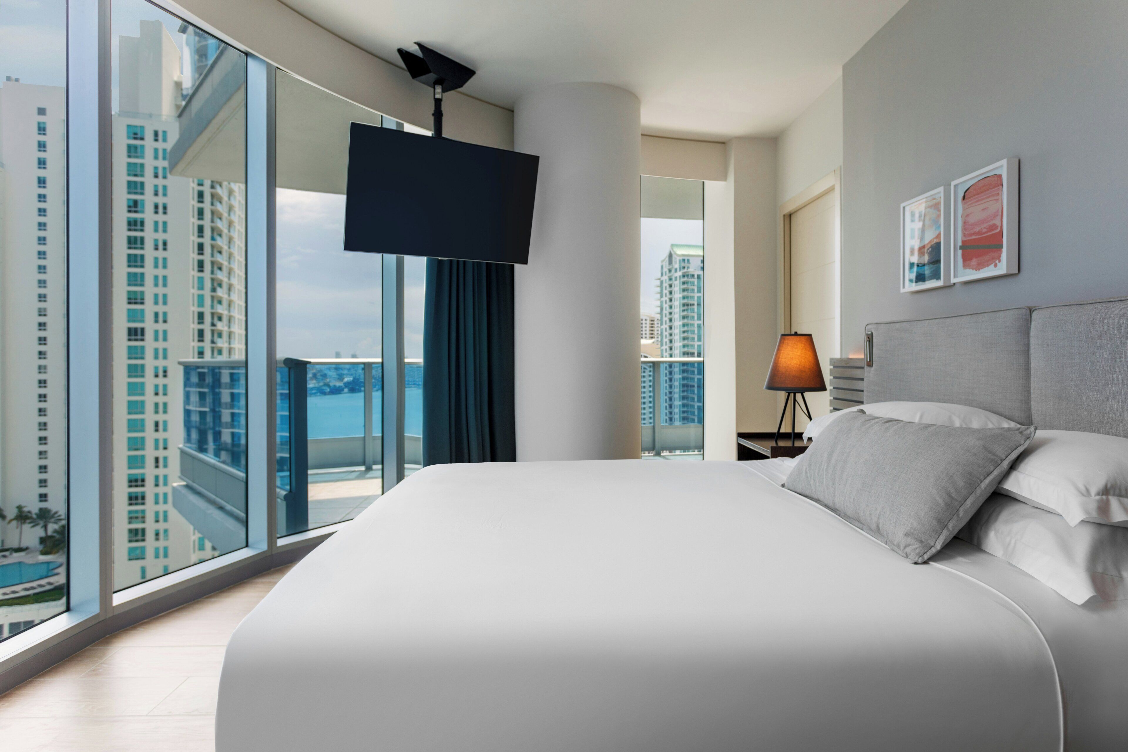 A modern hotel room with a large bed, a flat-screen TV, a city view, and a nightstand with a lamp.