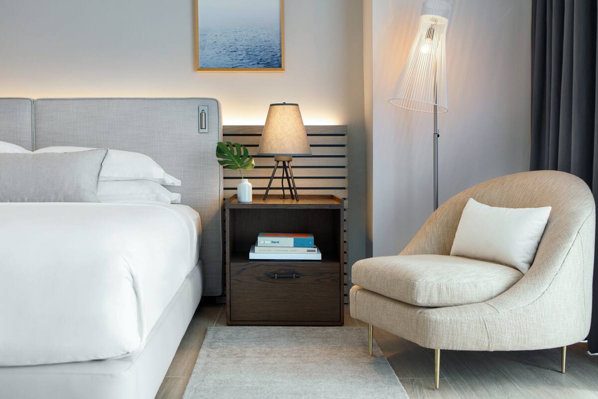 A modern hotel room with a bed, a nightstand with a lamp, a chair, and a bookshelf.