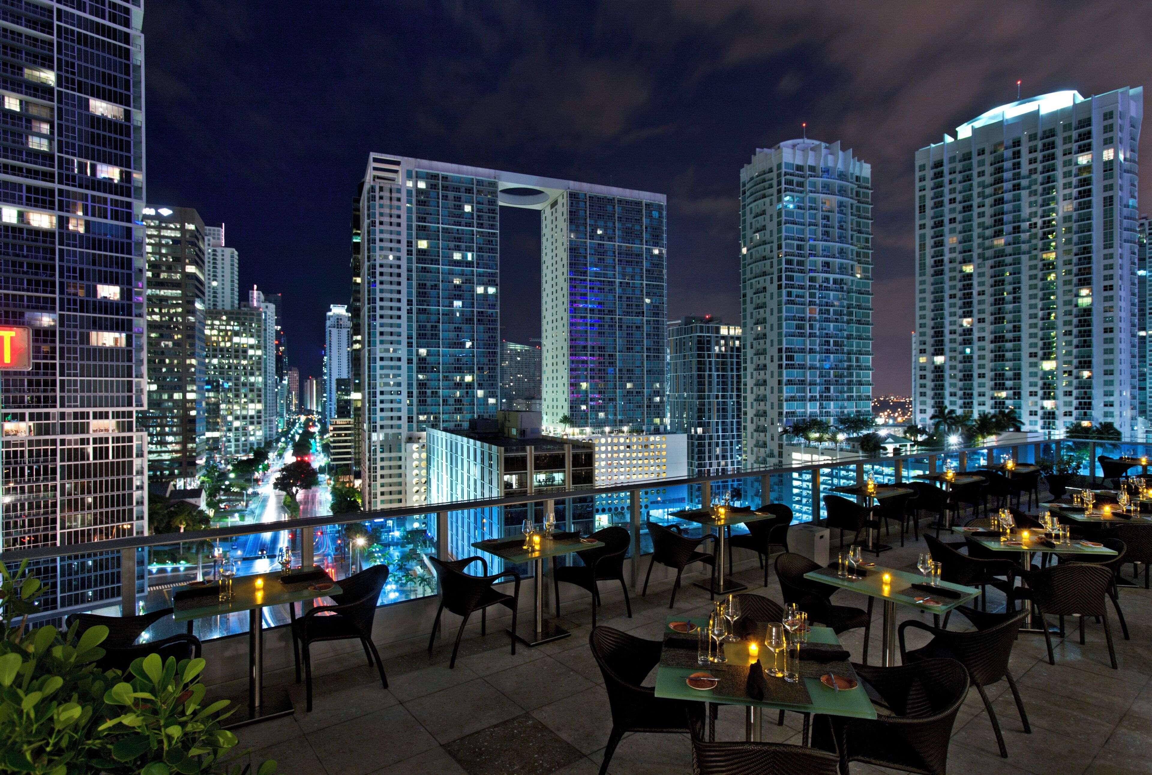 Un restaurante en la azotea con mesas y sillas, con vistas a un horizonte urbano nocturno.