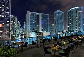 A rooftop restaurant with tables and chairs, overlooking a city skyline at night.