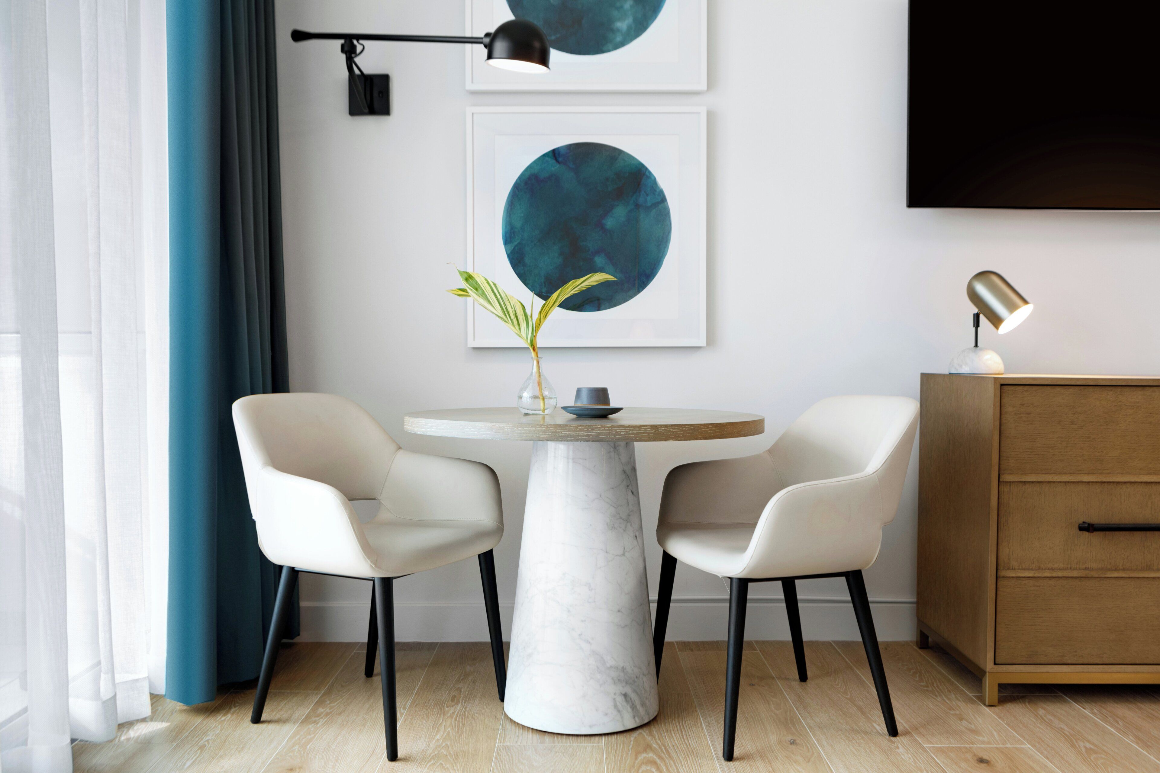 A modern dining area with a marble table, two chairs, a vase with a plant, and artwork on the wall.