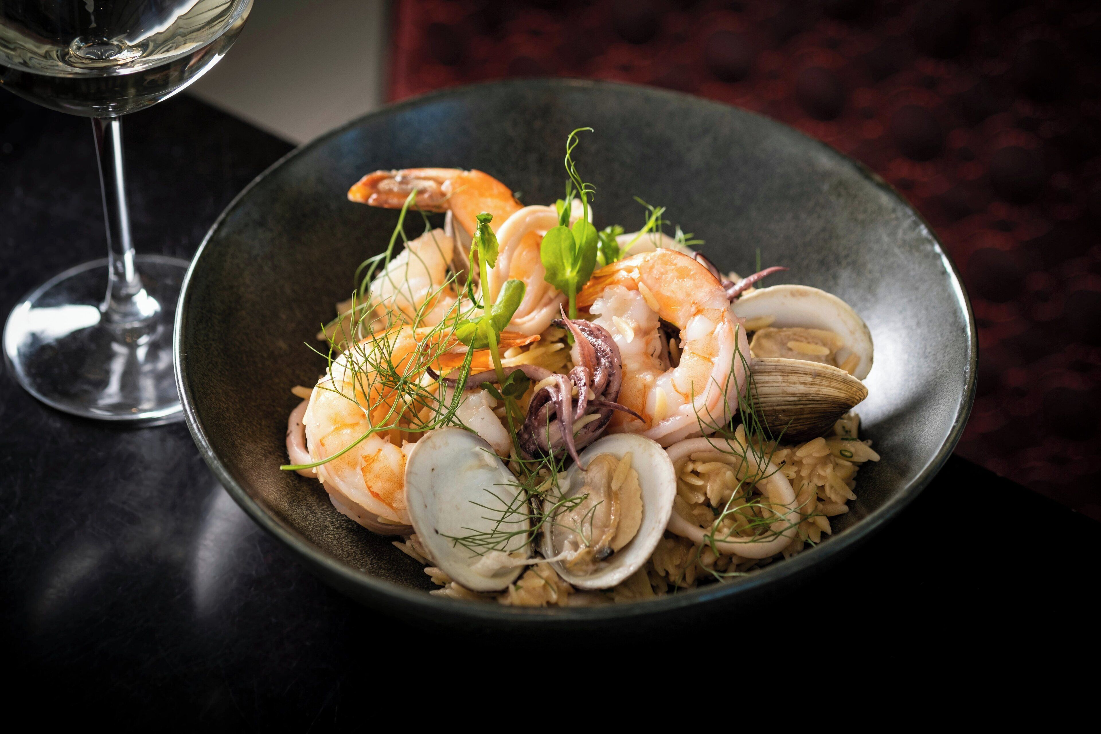 A bowl of seafood rice with clams, shrimp, and mussels, garnished with herbs, on a dark table.