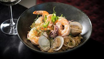 A bowl of seafood rice with clams, shrimp, and mussels, garnished with herbs, on a dark table.