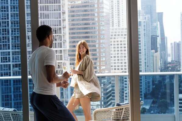 A man and a woman toasting with champagne glasses on a balcony with a cityscape view.