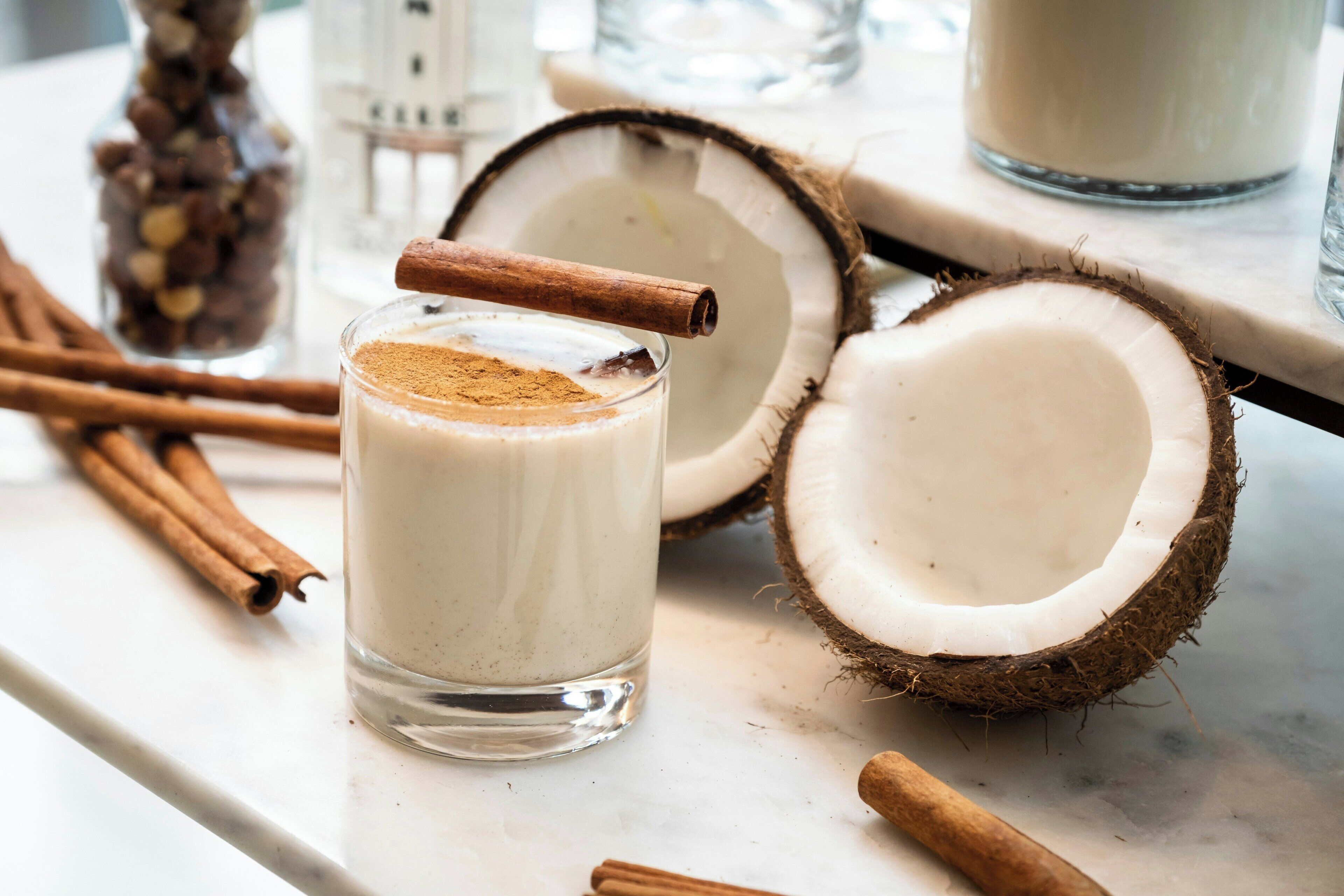 Un vaso de leche de coco con canela sobre una superficie de mármol, rodeado de cocos partidos y ramas de canela.