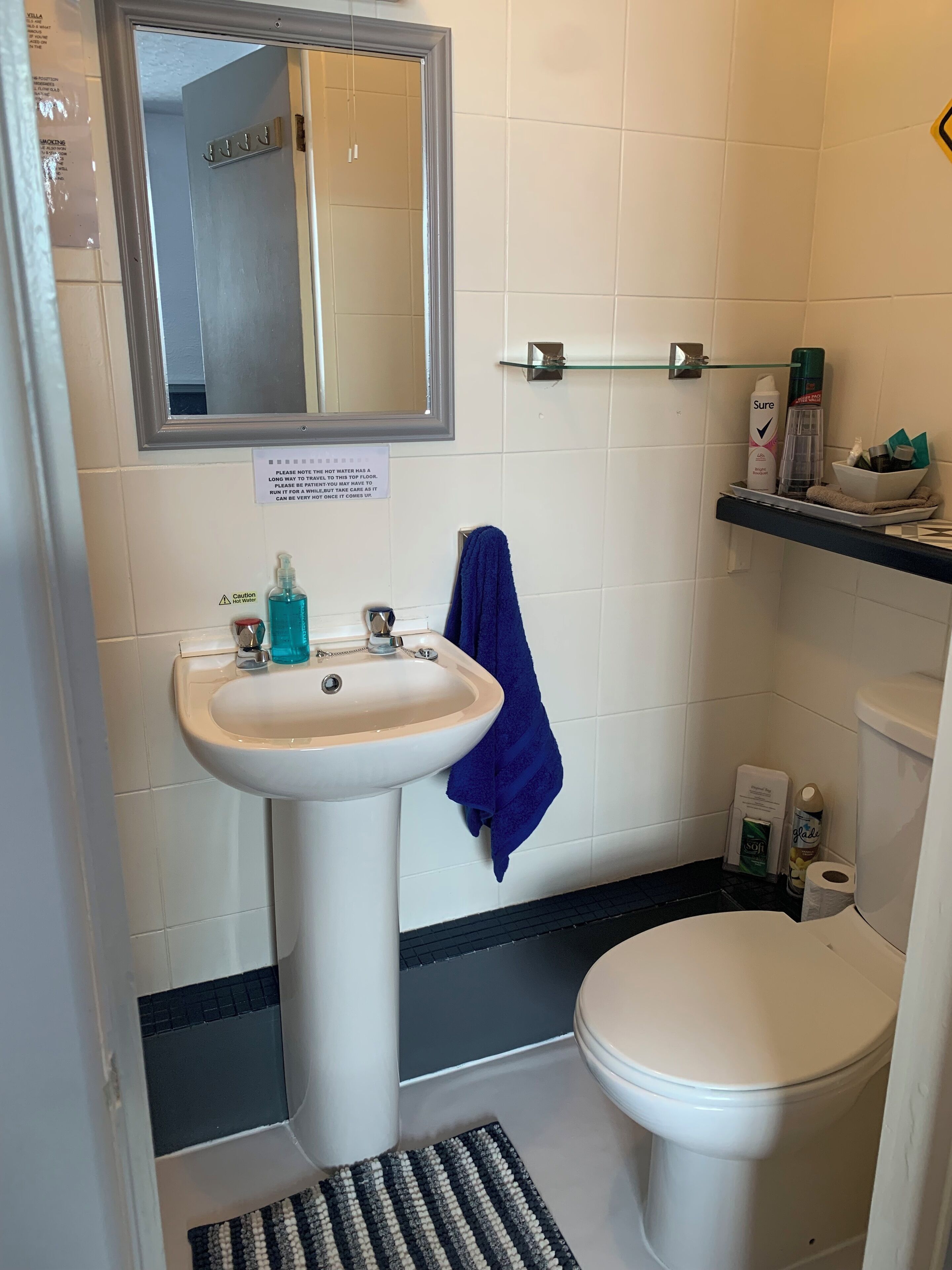 A bathroom with a sink, mirror, and toilet. There is a caution sign about hot water, a towel rack, and toiletries on a shelf.