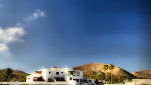 A modern white building with multiple floors and a flat roof, surrounded by palm trees and a clear blue sky. - Hotel Rural Finca de la Florida (San Bartolome)