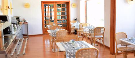Un espace repas avec des tables et des chaises en bois, un coin cuisine équipé d’appareils électroménagers et une grande fenêtre qui laisse entrer la lumière naturelle.