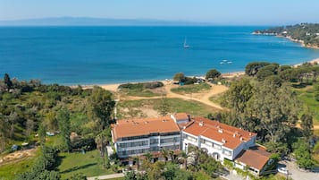 A large, multi-story building with a red-tiled roof, surrounded by lush greenery and a beach.