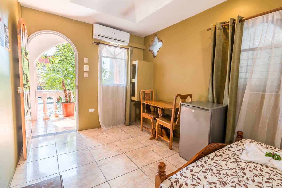 Une chambre comprenant un lit, un bureau, une chaise, un petit réfrigérateur et un appareil de climatisation. Il y a une fenêtre avec des rideaux et une vue sur la verdure à l’extérieur.