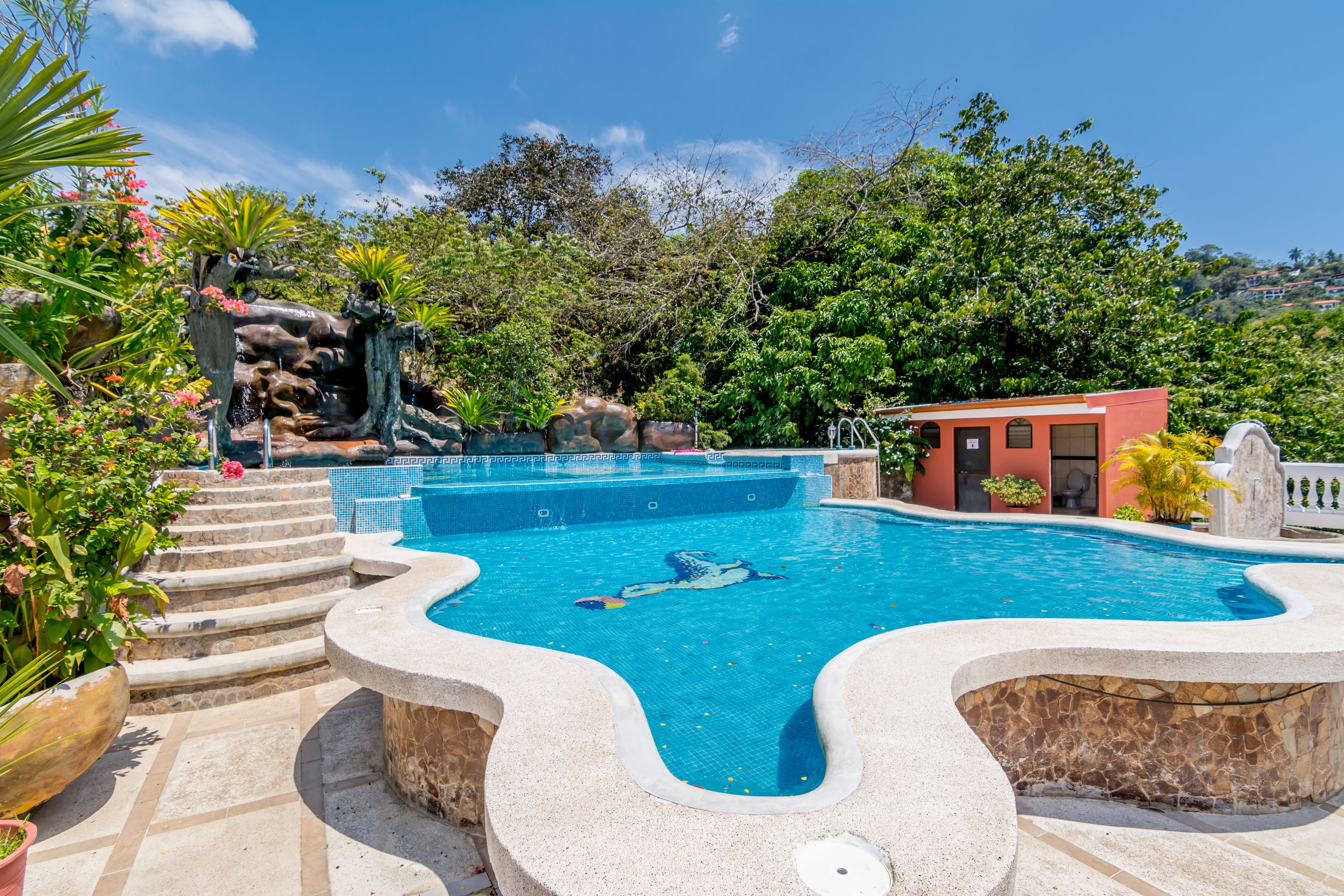 A swimming pool with a waterfall feature, surrounded by lush greenery and a small building.