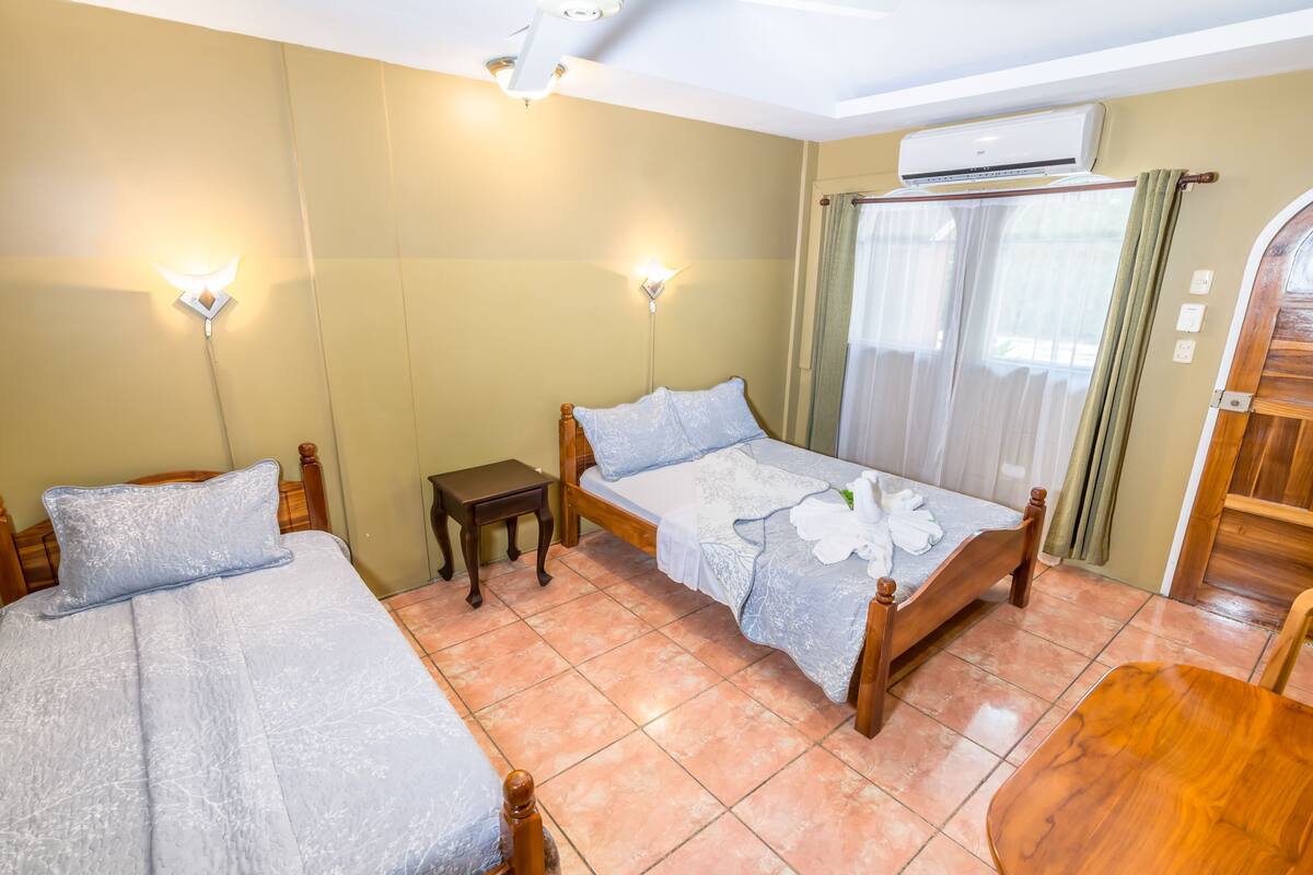 Une chambre d’hôtel avec deux lits, une petite table, une chaise en bois, un ventilateur de plafond et un appareil de climatisation.