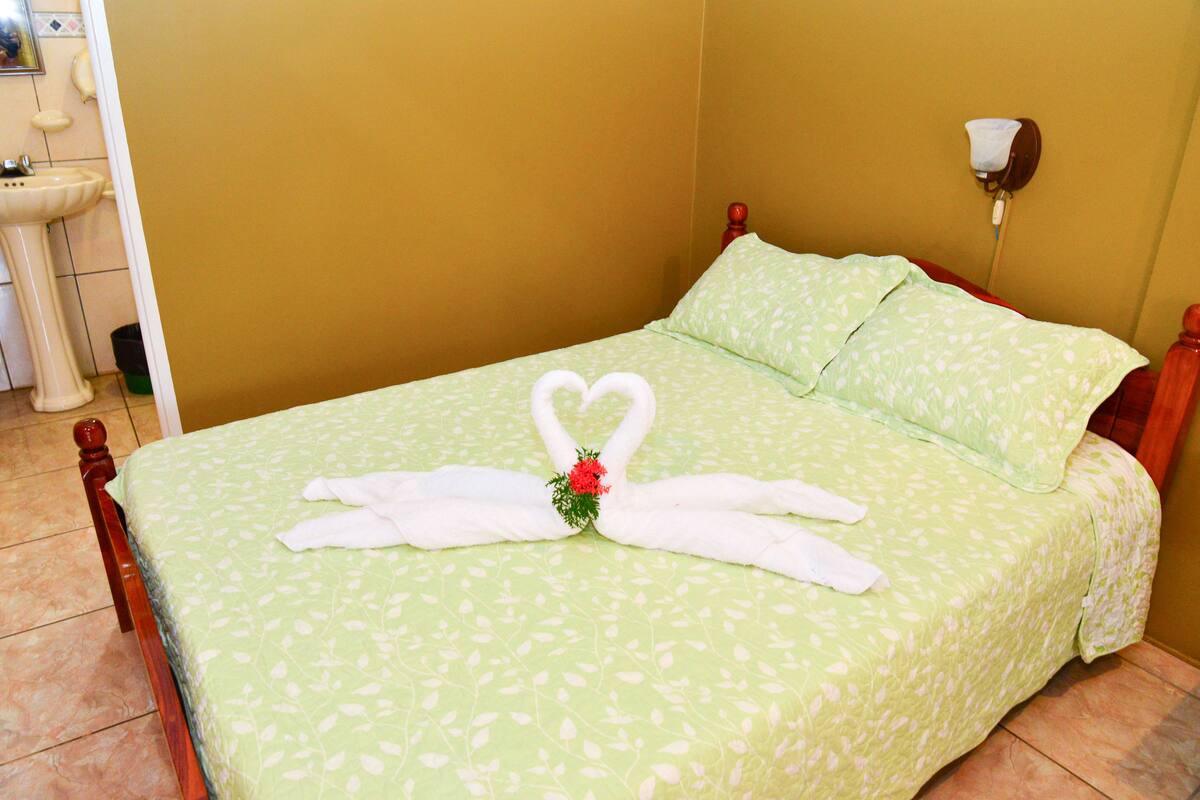 Un lit bien fait, recouvert d’une couvre-lit à motifs floraux verts, et agrémenté d’une serviette pliée en forme de cœur.