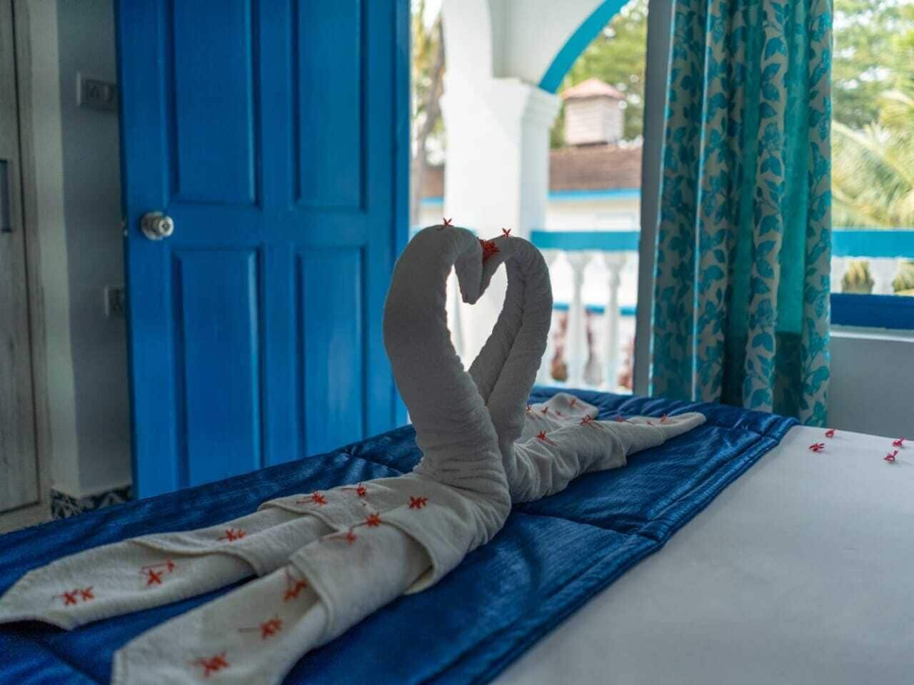 Una cama con una toalla dispuesta en forma de corazón, una puerta azul y una ventana con cortinas.