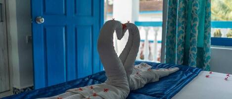 Una cama con una toalla dispuesta en forma de corazón, una puerta azul y una ventana con cortinas.