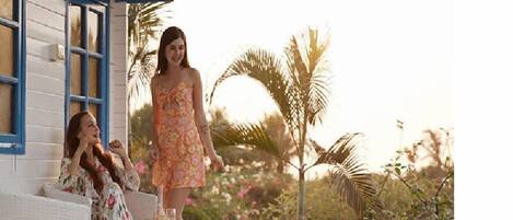 A woman in a floral dress standing on a porch with a white building and blue shutters in the background.