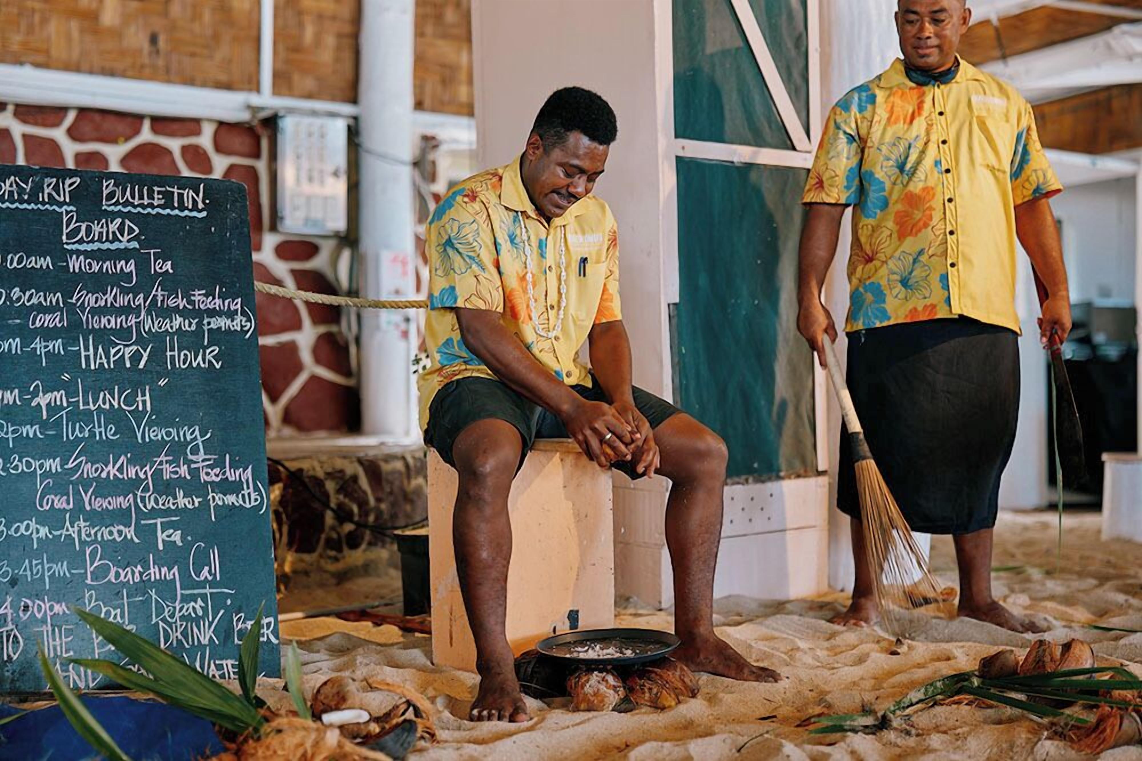 A man sitting on a stool at a beachside restaurant with a chalkboard menu listing breakfast and lunch items.