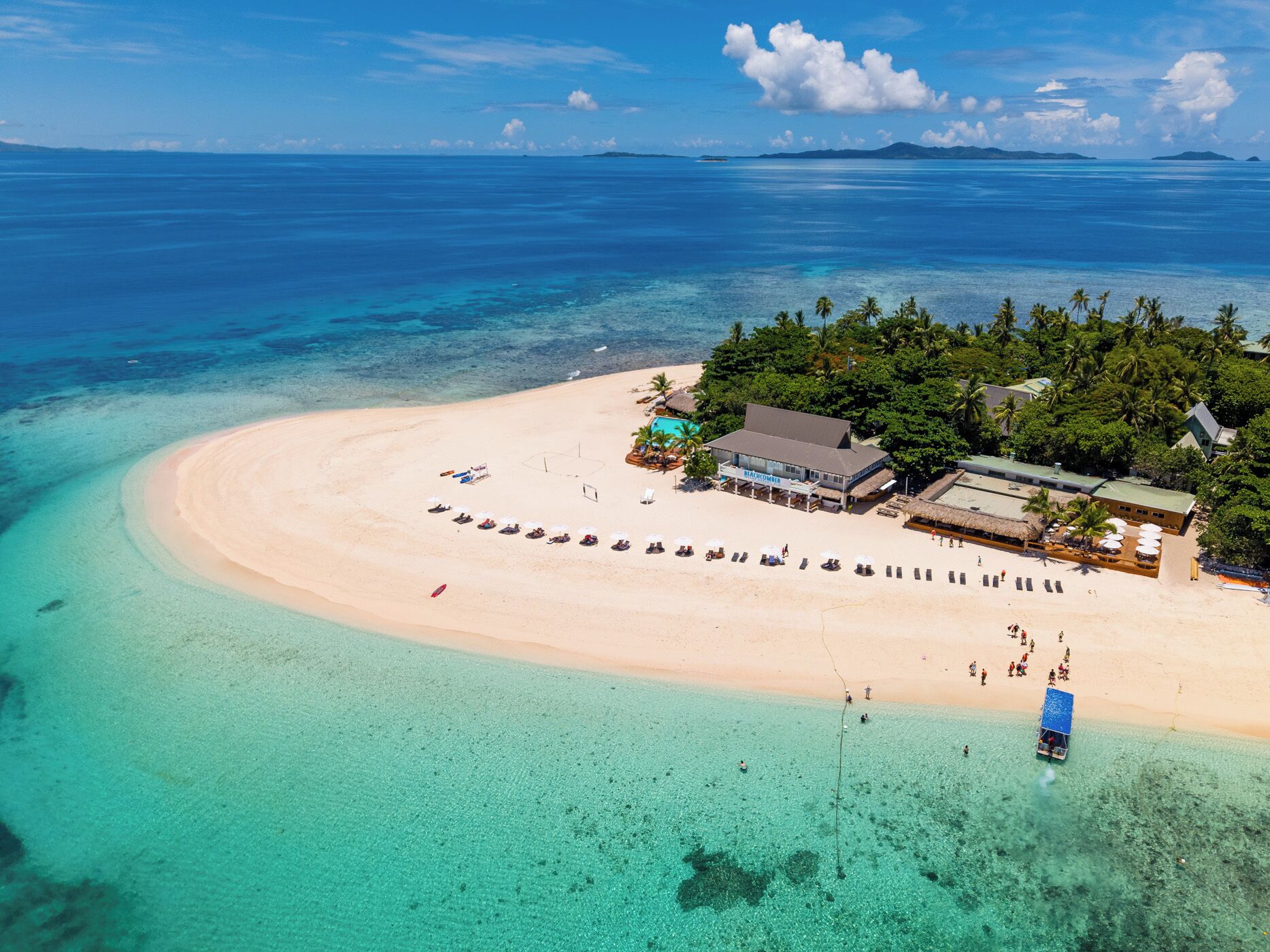 Luftaufnahme eines Strandresorts mit einem großen Gebäude, einem Schwimmbad und einem Strand mit Liegestühlen.