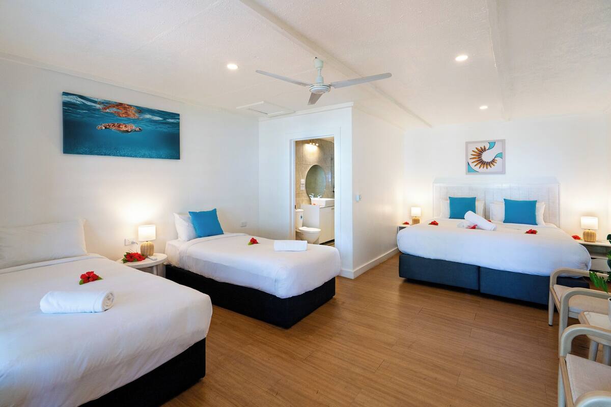 Une chambre d’hôtel avec trois lits, un ventilateur de plafond et un tableau représentant une tortue de mer.