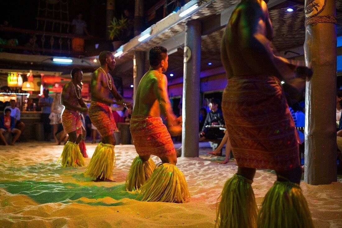 A group of people performing a traditional dance on a sandy surface with a bar and people in the background.