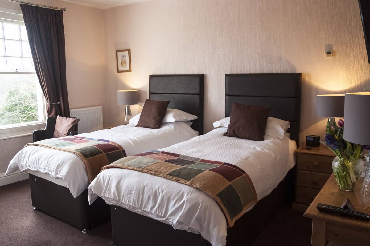A hotel room with two beds, a window with curtains, bedside lamps, a nightstand with a vase of flowers, and a remote control.
