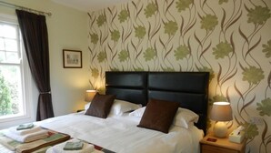 A neatly made bed with a headboard, two bedside lamps, a wooden nightstand, and a window with curtains. - Overcombe House (Yelverton)