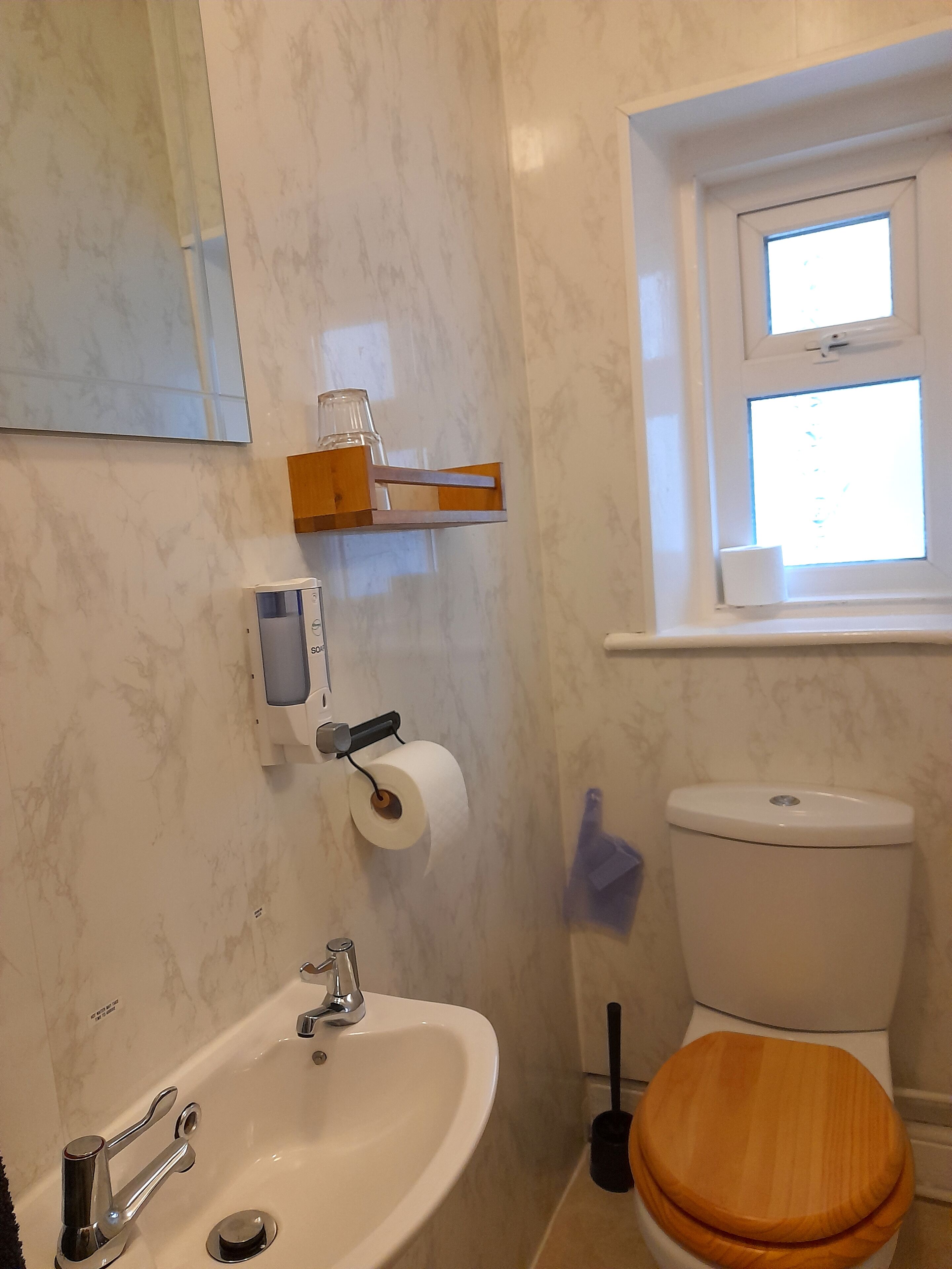 A bathroom with a white sink, a toilet with a wooden seat, a window above the toilet, and a mirror on the wall.