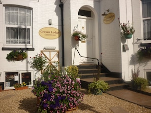 Een wit gebouw met een bord waarop 'Crown Lodge Guest House' staat en een bord met 'Geen kamers beschikbaar'. Er staan bloembakken onder de ramen en een kleine tuin met bloemen.