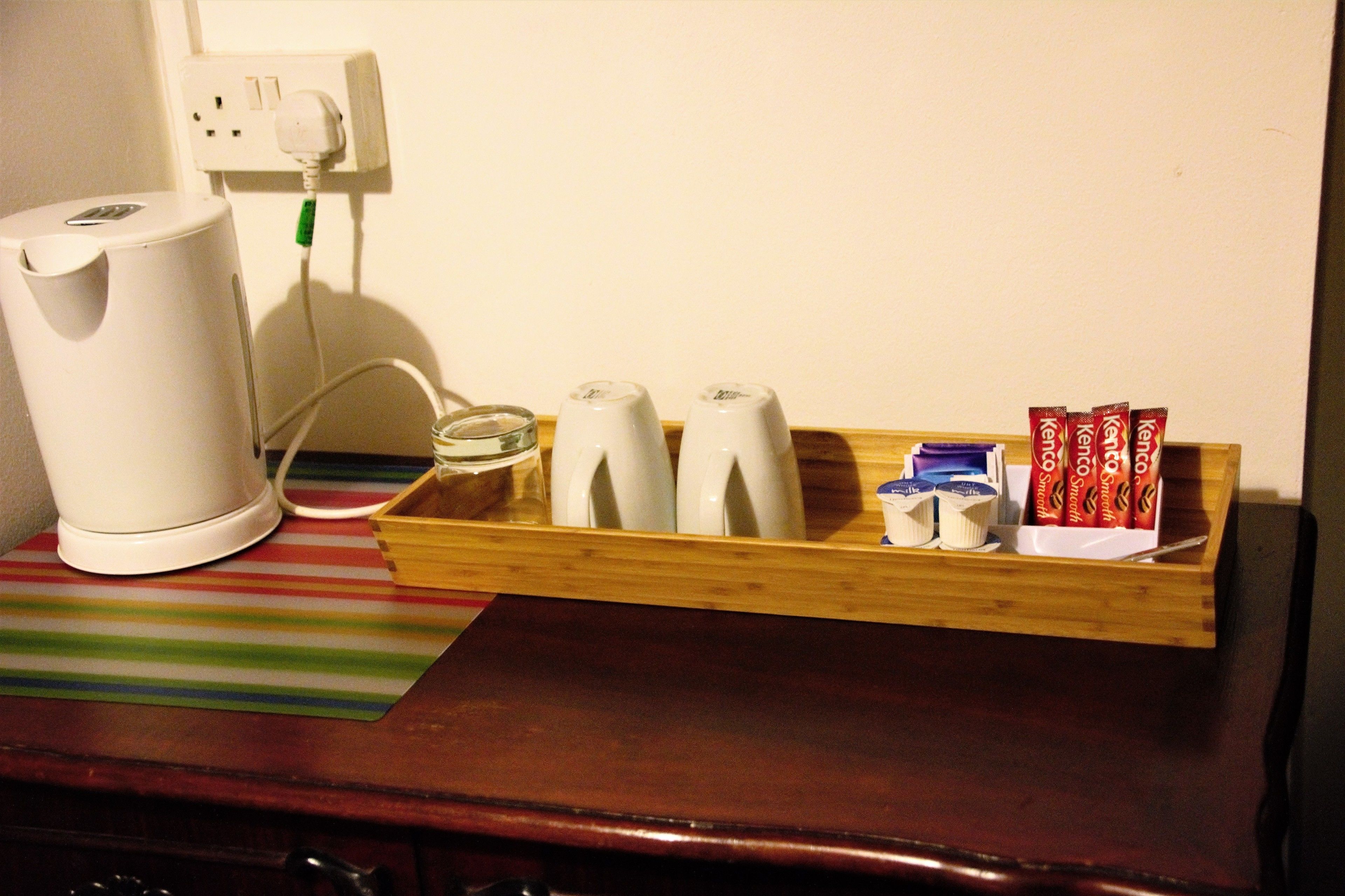 A wooden tray with a white kettle, a glass, and some packaged snacks on a table.