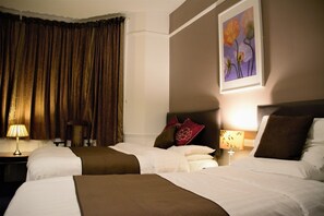 Une chambre d’hôtel avec deux lits, une grande fenêtre avec des rideaux, un tableau floral encadré et une table de chevet avec une lampe.