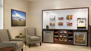 Un área de espera moderna con dos sillones grises, una mesita redonda pequeña, un refrigerador lleno de aperitivos, un enfriador de bebidas y un estante de pared con diversos productos envasados.