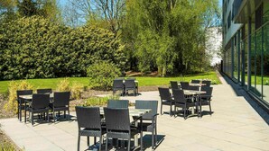 An outdoor patio with tables and chairs, surrounded by greenery and a building with large windows. - ibis Styles Crewe (Crewe)