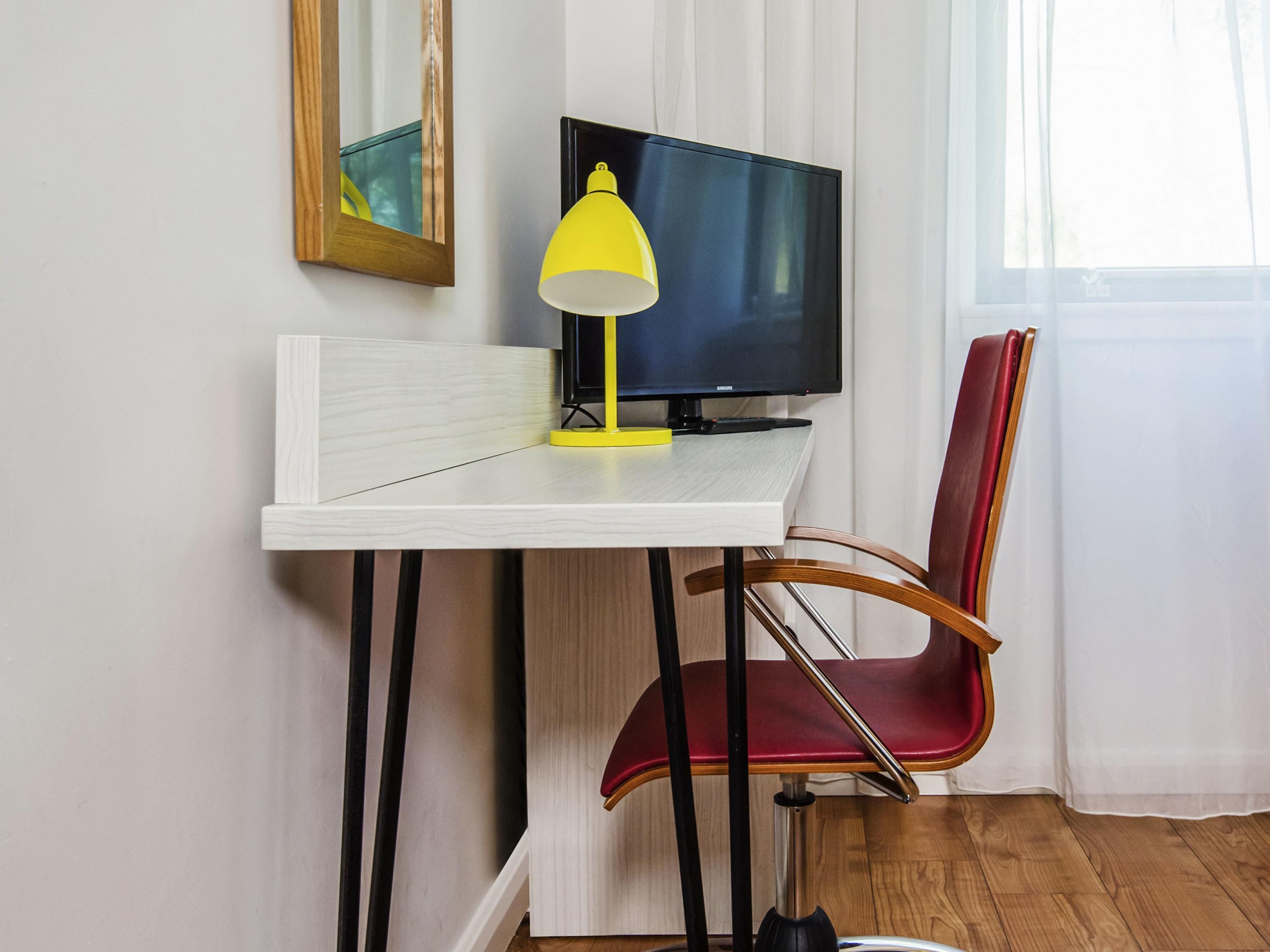 Quarto com escrivaninha branca, abajur amarelo, cadeira vermelha, televisão preta e janela com cortinas brancas.